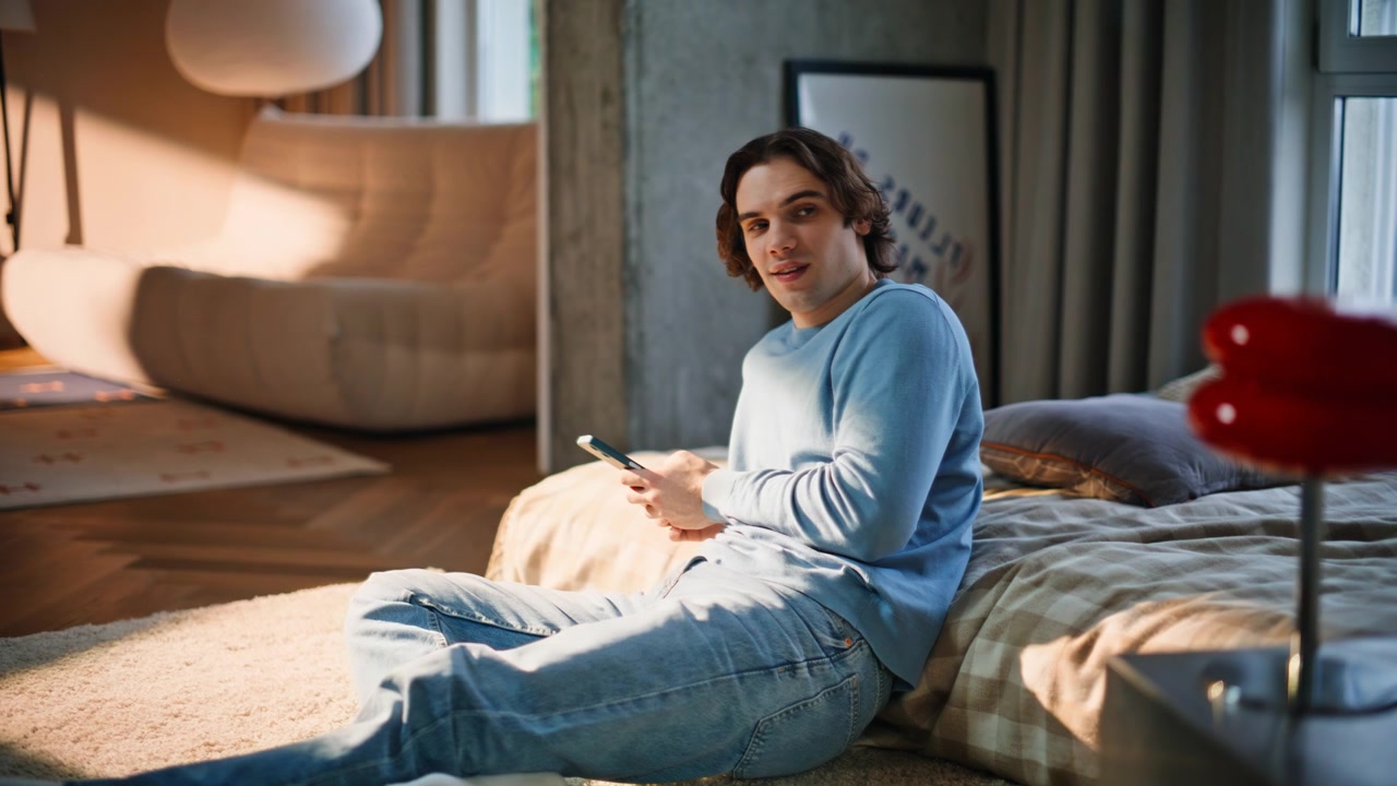 Uomo che si rilassa leggendo un messaggio sullo smartphone in una camera da letto illuminata dal sole. Ragazzo seduto per terra.