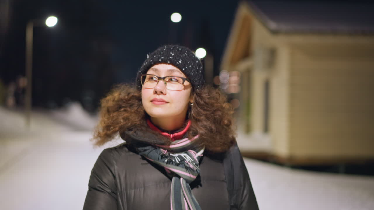 Woman in warm winter jacket and knitted hat standing outdoors on snowy evening street, scarf around neck, glasses reflecting light, looking up with peaceful expression, by soft glowing streetlights