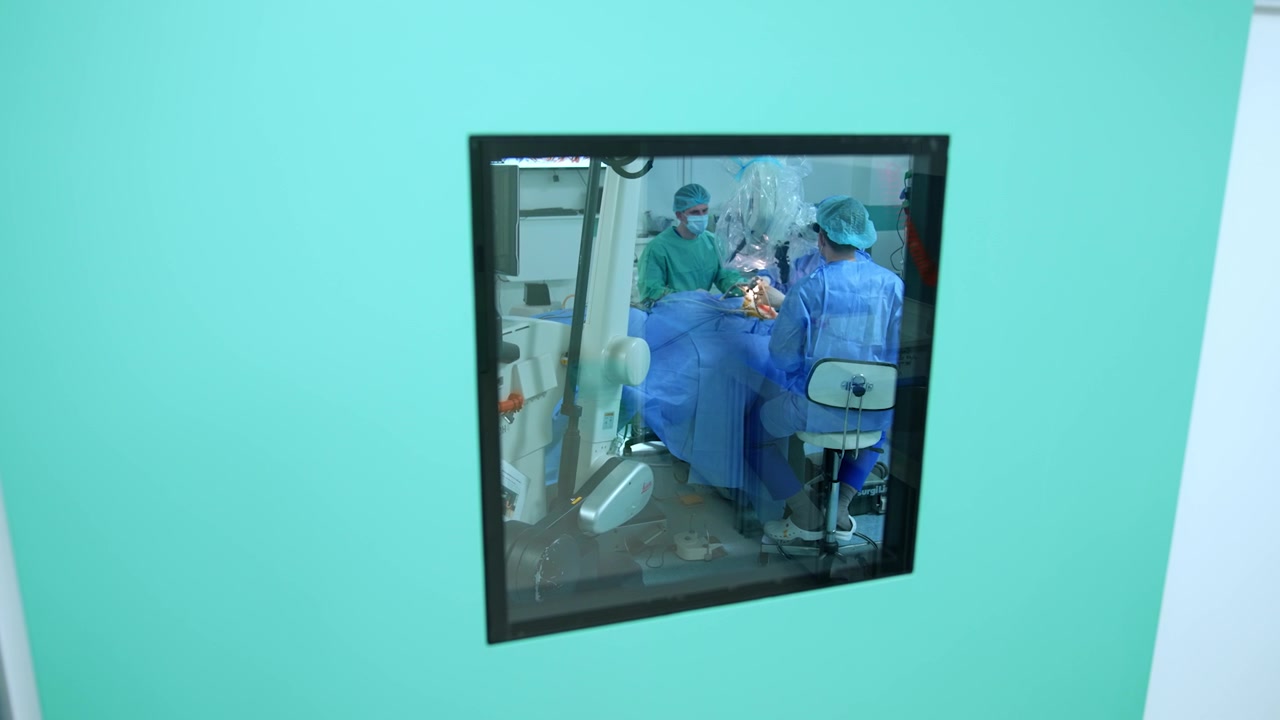 Square window in the surgery room door. Two neurosurgeons sit at the operating table performing operation.