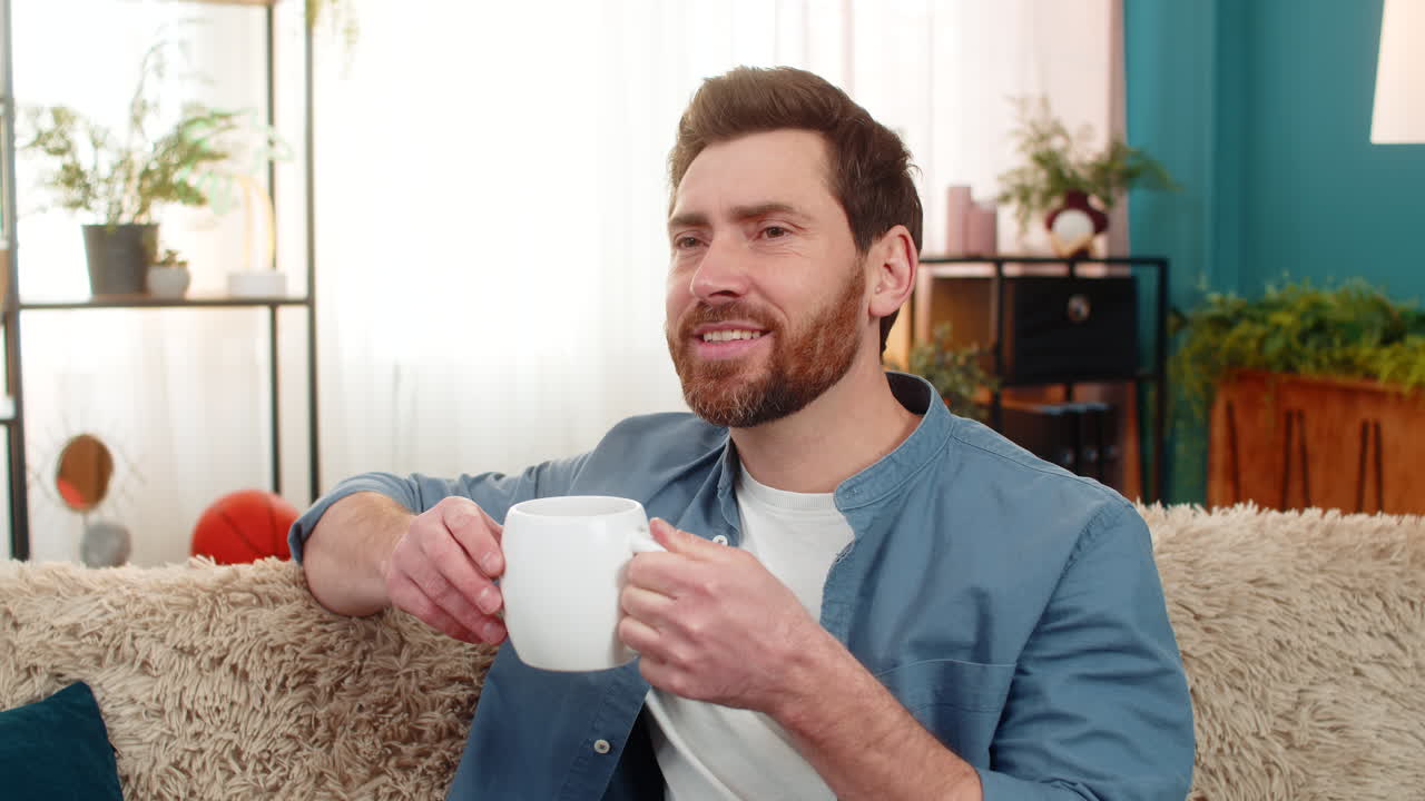 Young man sitting on sofa holding a cup drinking tea gazing into distance enjoying the moment