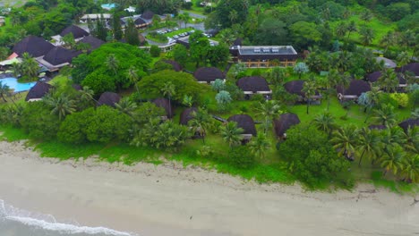 From-high-above,-the-drone-surveys-the-Tieti-hotel,-standing-out-like-a-pearl-amidst-the-crystal-clear-waters-and-golden-sands-of-Poindimie,-in-the-exotic-island-paradise-of-New-Caledonia