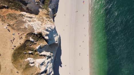 aerial top down flight over sandy beach along cliff coastline during sunny day - people walking along shore of sea - etretat, france