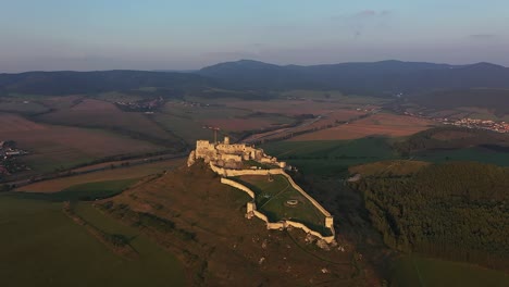 a drone circles majestic spiš castle at sunset, golden light illuminating the fortress, with blue-grey clouds and vast green meadows all around