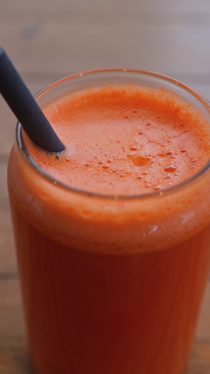 Primo piano di un succo di carota e arancia in un bicchiere con una cannuccia nera su un tavolo di legno. Verticale