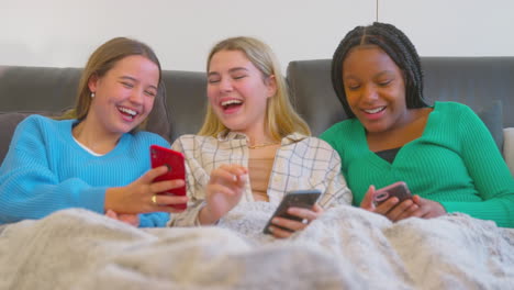 group of multi-cultural teenage girl friends snuggled under blanket looking at mobile phones at home