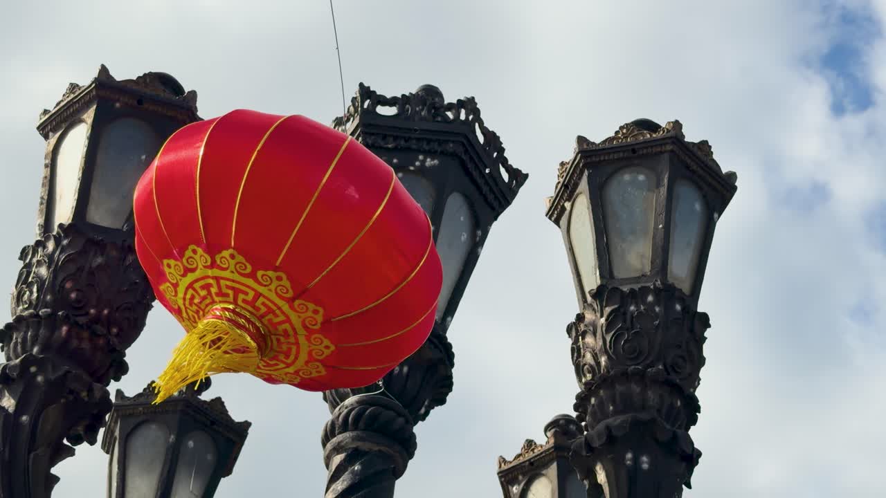 Cảnh quay góc thấp một chiếc đèn lồng đỏ đung đưa bên cạnh những cột đèn cổ điển trang trí công phu, trước nền trời nhiều mây