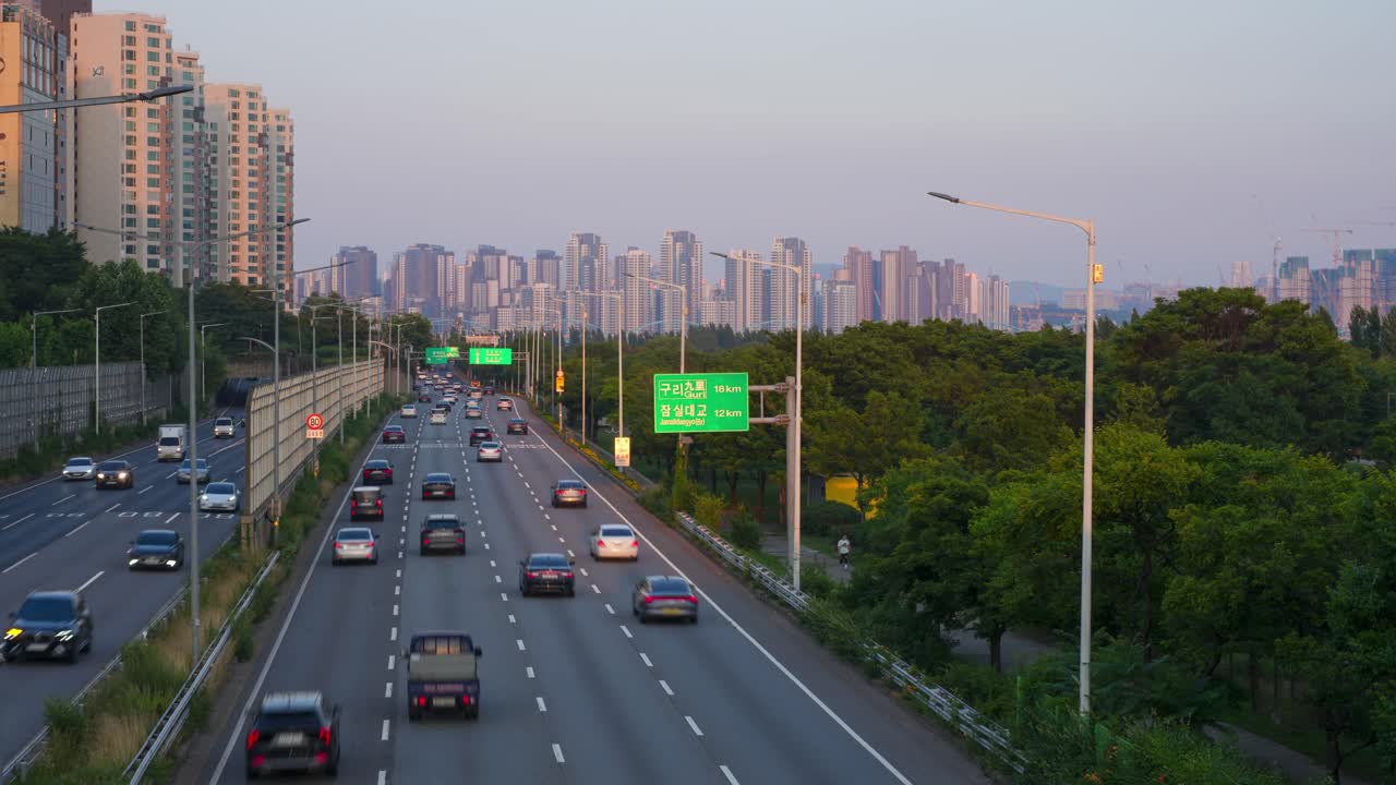 Time-lapse of busy highway in Seoul at sunset, with cars moving through cityscape flanked by high-rise buildings and lush trees