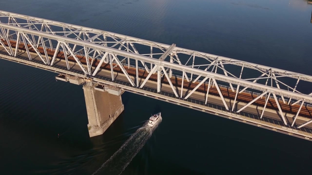 Aerial footage of a boat traveling through the blue waters of the Little Belt Strait, passing beneath the historic Old Little Belt Bridge in Middelfart, Denmark.