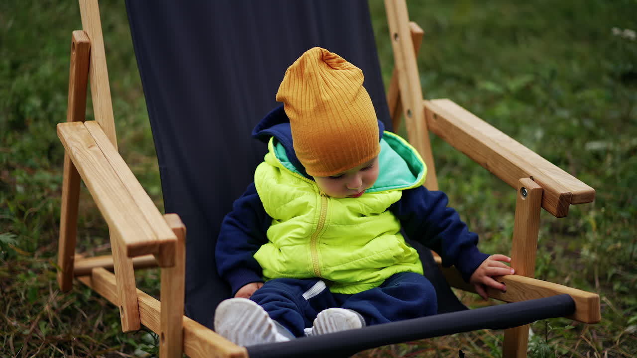 Caucasian toddler wearing warm clothes and cap sitting in garden chair. Baby looking around cautiously sitting peacefully. Close up. Blurred nature backdrop.