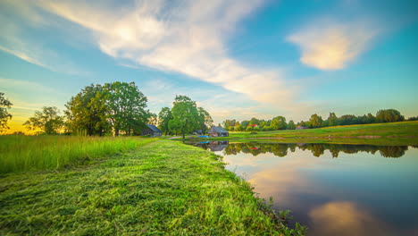 Static-shot-of-sun-going-up-in-timelapse-over-a-wooden-cabin-beside-beside-a-small-lake-along-rural-landscape