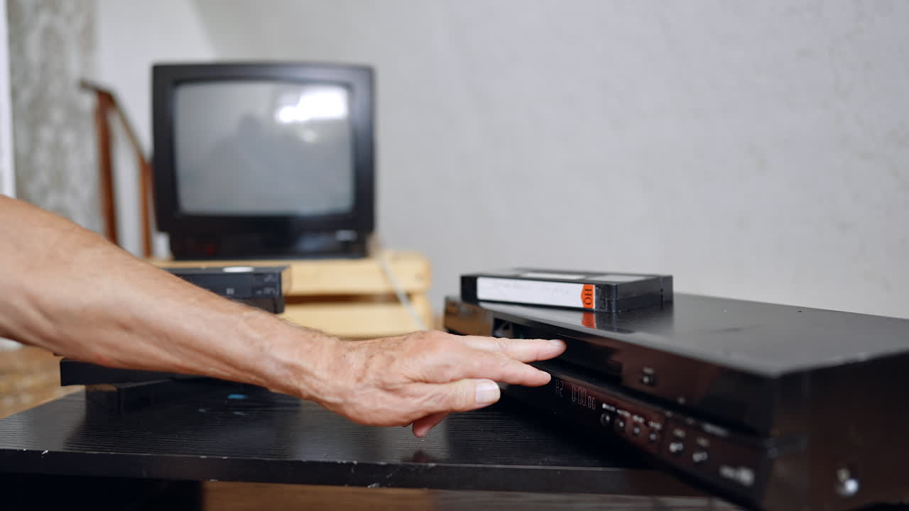 Hand holding VHS cassette by TV. A person holds a VHS tape near a vintage television and VCR inside a modest room