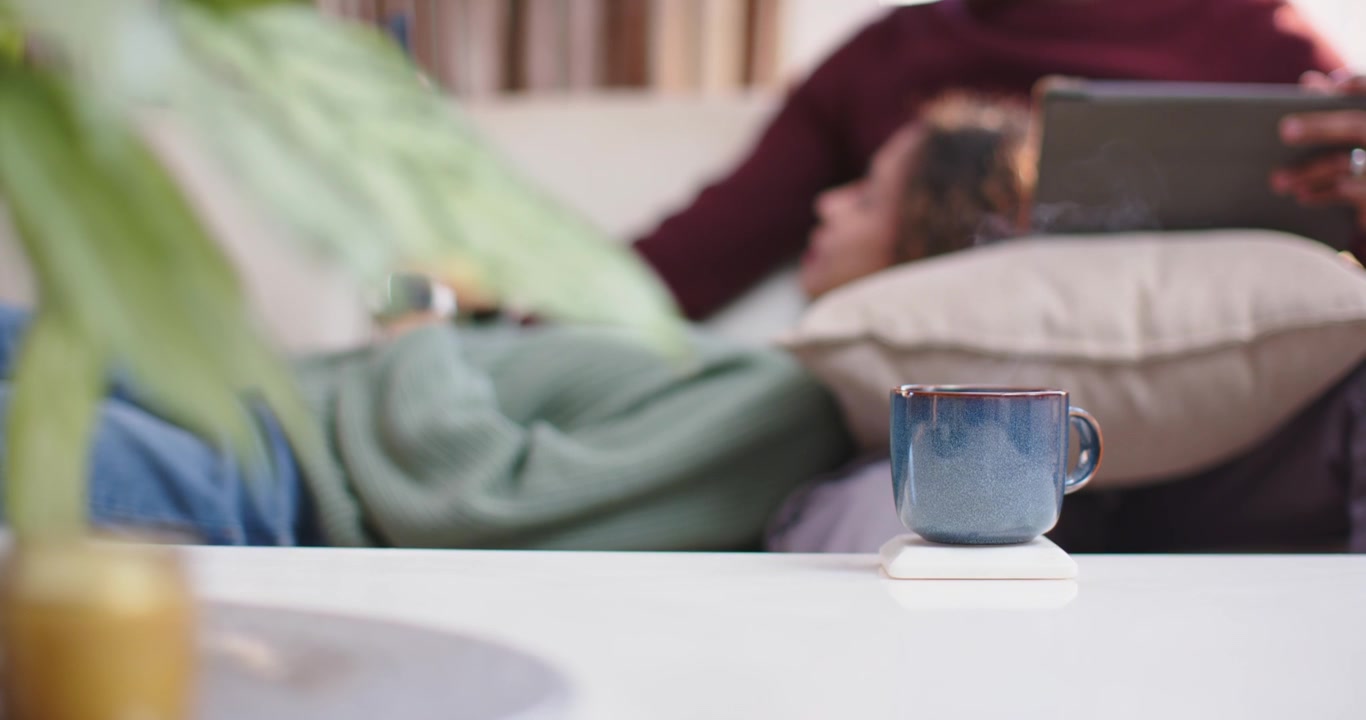 カップルがソファにもたれかかり、コーヒーテーブルのコースターの上には青いマグカップ。男性は読書しながらタブレットをスクロールしている。