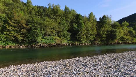 Revealing-running-waters-of-lake-next-to-stones-and-trees,-parallax-drone-shot