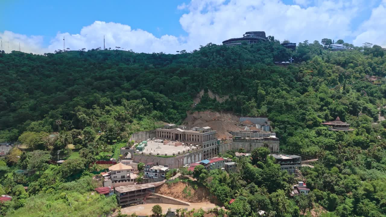 Elevated drone shot of a neoclassical estate on a forested hillside in the Philippines, showing lush mountains, village homes, and tropical greenery under blue skies