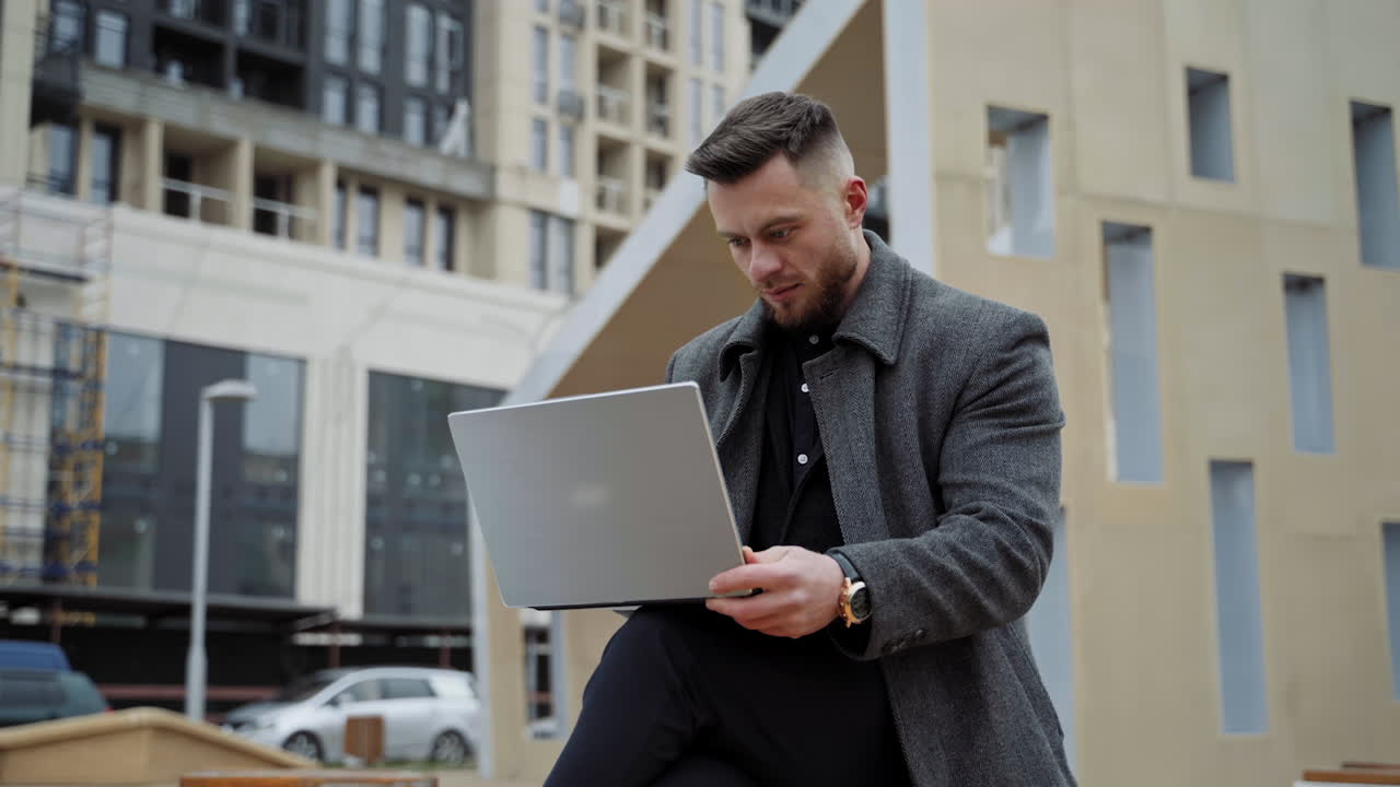 Young man with a laptop on fresh air. Handsome young man in a coat sitting in the street and working on a modern device in the city. Distance work.