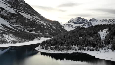 Aerial-flyover-over-shores-lake-Oeschinensee-in-Kandersteg,-Switzerland-on-a-cold-winter-day-with-snow-covered-trees-and-reflection-of-peaks