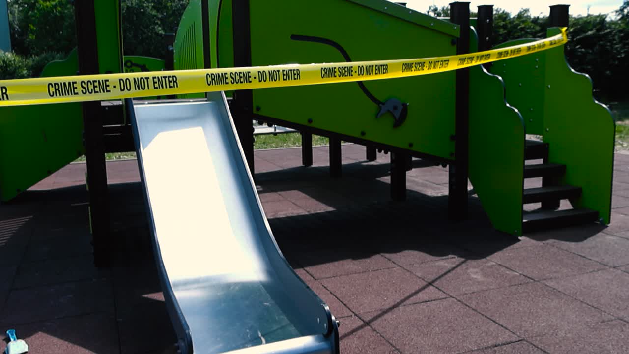 Vibrant colored footage of a yellow Crime Scene Do Not Enter tape in front of a childrens playground slide during a sunny summer day. The tape is moving slowly in the wind and no people are visible.