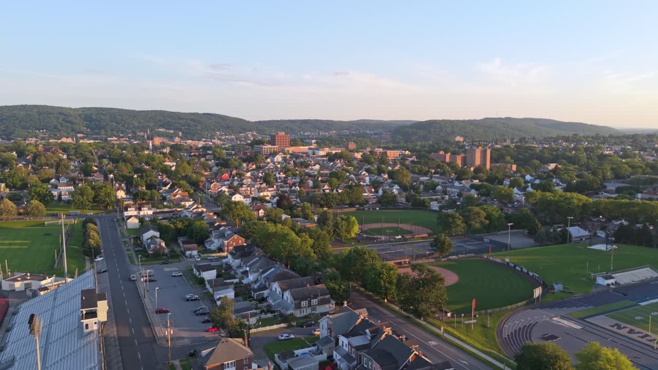 Aerial flyover of Bethlehem, Pennsylvania with sports stadiums, houses, and distant hills under warm sunset light. Dolly in