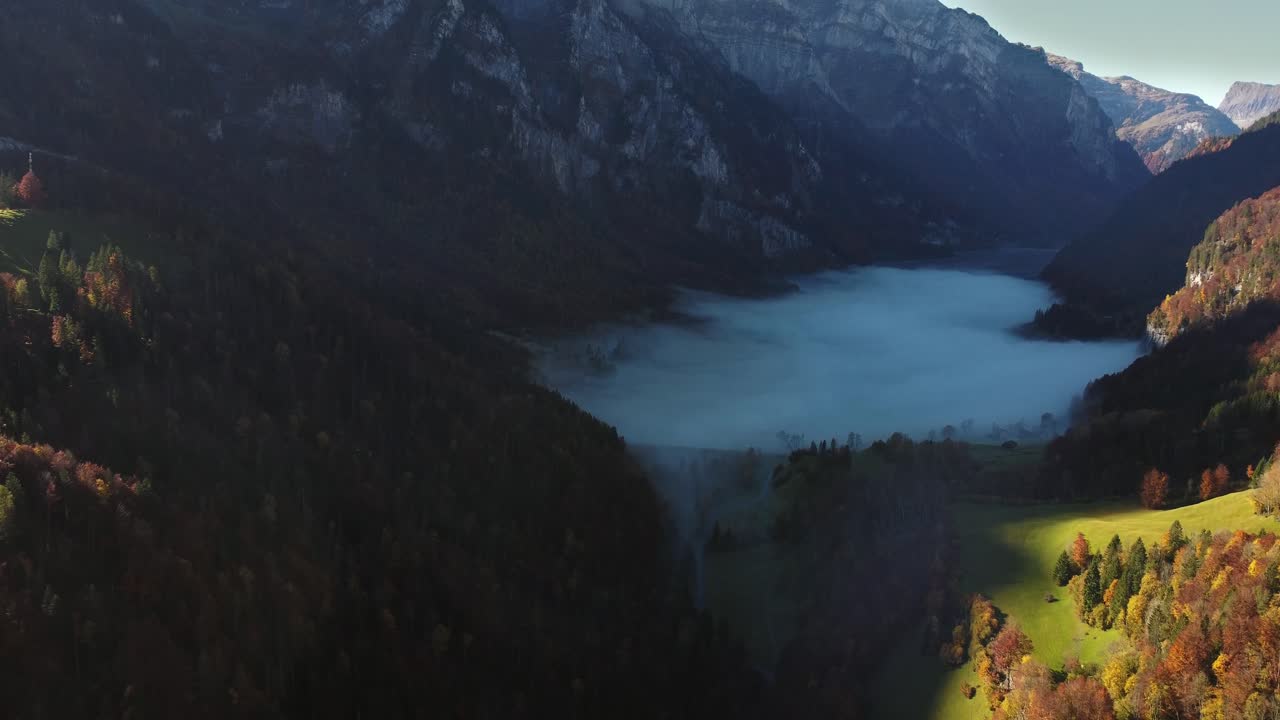 Riprese aeree con drone del lago Klöntalersee coperto dalla nebbia mattutina e circondato da foreste autunnali. La luce del sole illumina i pendii e le scogliere colorati delle Alpi svizzere. Scena naturale calma e cinematografica