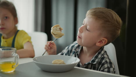 young boy and girl sitting at dining table eating dumplings, boy taking big bite while girl looks curiously toward background, bright home setting with juice glasses and cozy casual atmosphere