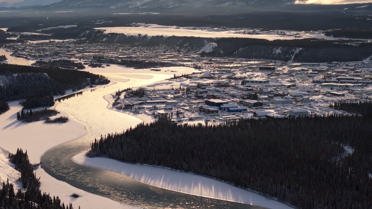 A smooth aerial tilt-up establishing shot at sunset, capturing Downtown Whitehorse, the winding Yukon River, and the rugged, untouched landscape surrounding the city in this peaceful northern setting.