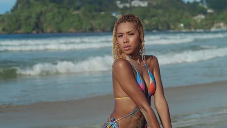 At-Las-Cuevas-Beach-in-Trinidad,-a-sunny-Caribbean-day-sees-a-young-girl-with-curly-hair-in-a-bikini-by-the-waves-kneel-in-the-sand-at-the-shoreline