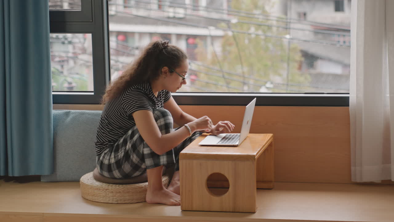 Young woman working laptop hotel room, seated on floor cushion by large window with city view, casual dress, typing emails and checking messages, sipping coffee between tasks, focused expression