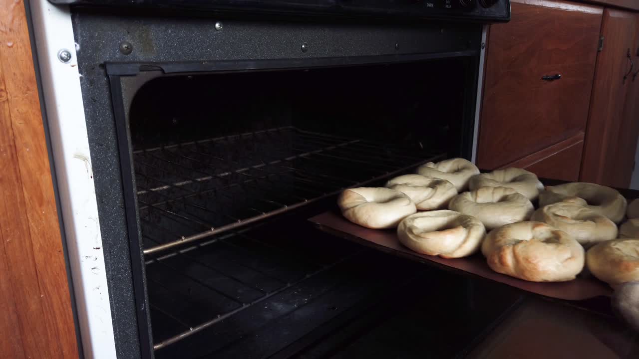 Manos con manoplas de horno retiran una bandeja de bagels recién horneados de un horno casero.