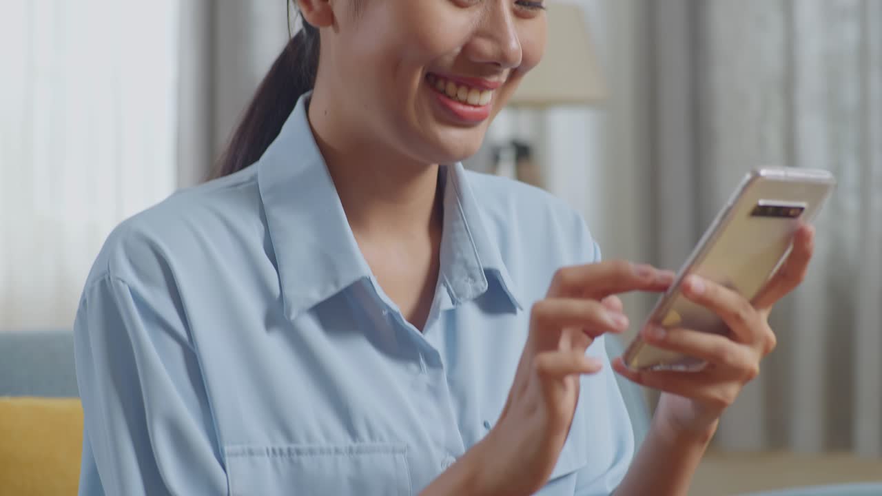 아시안 여성이 거실의 소파에서 스마트폰을 사용하면서 커피를 마시고 있습니다.