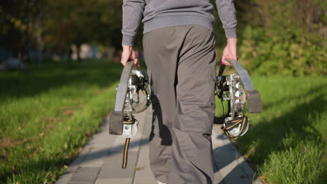 vista posterior de una persona con pantalones sueltos de color gris oscuro y sudadera que se aleja por un camino pavimentado de color gris claro, llevando pilotes metálicos plateados y negros con componentes mecánicos visibles, bordeados por hierba verde