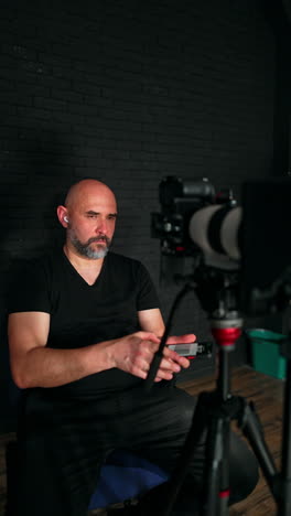 bald caucasian man wearing black clothes sits behind the camera. cameraman takes and uses his phone. studio backstage. vertical video.