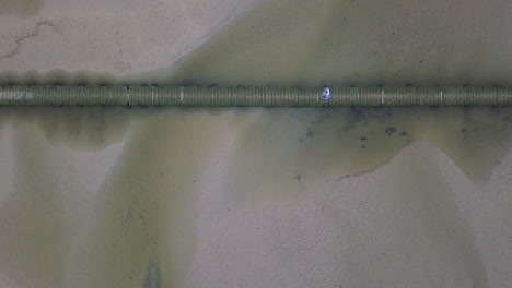 Man-Walking-On-Wooden-Bridge-Across-Sandy-River-Gannel-In-Newquay,-UK---aerial-top-down