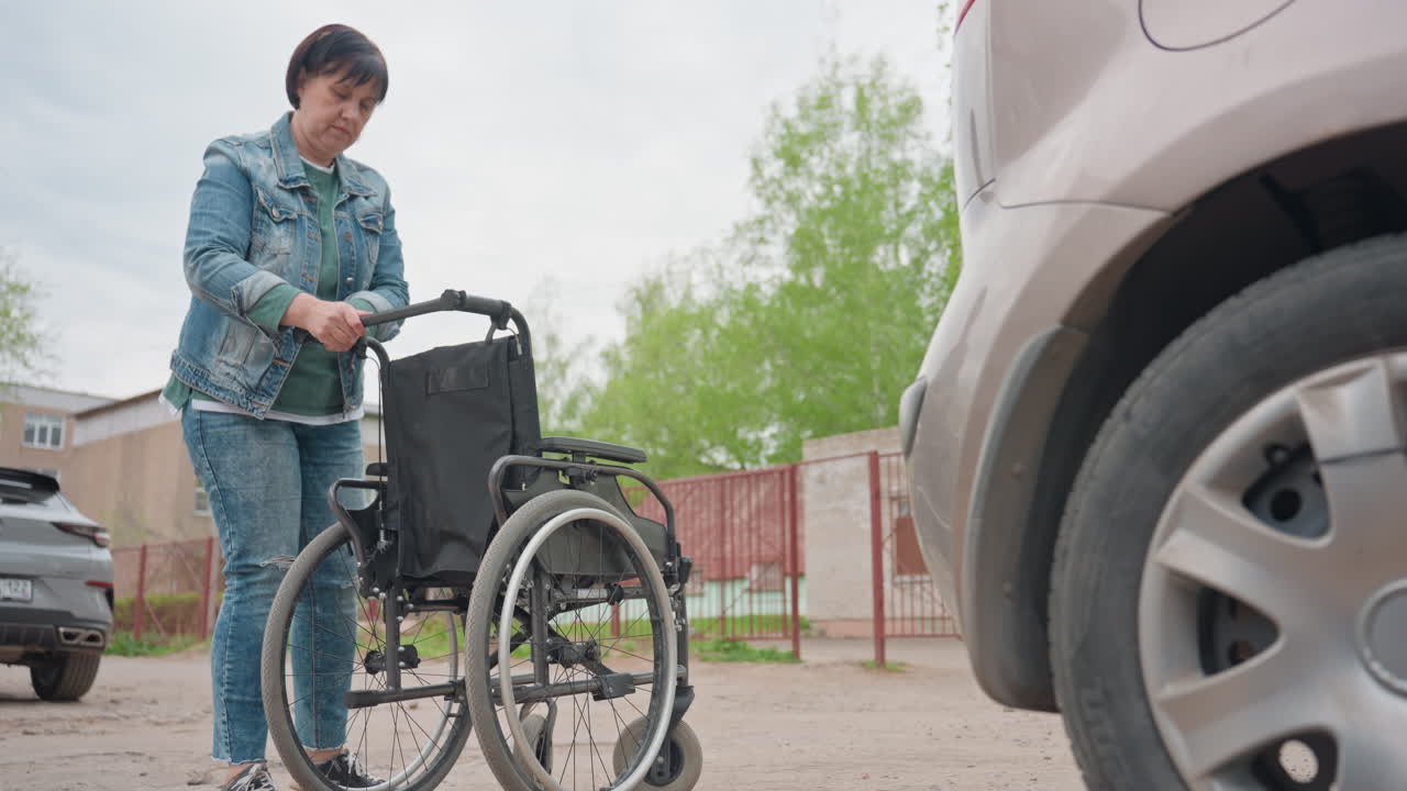 Woman Prepares Wheelchair Beside Parked Car, Denim Jacketed Adult Adjusts Manual Wheelchair In Suburban Parking Lot, Residential Buildings In Background, CloseUp On Wheel And Vehicle Tire, Determined
