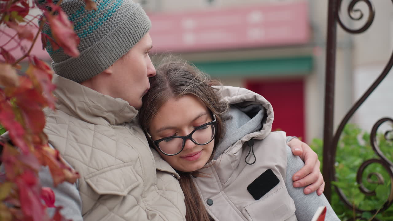 Side view of lovers wearing matching winter outfits as man kisses woman head affectionately while she smiles warmly, leaning on him with comfort and love, residential building visible in soft background