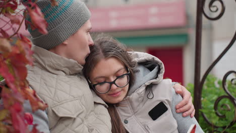 side view of lovers wearing matching winter outfits as man kisses woman head affectionately while she smiles warmly, leaning on him with comfort and love, residential building visible in soft background