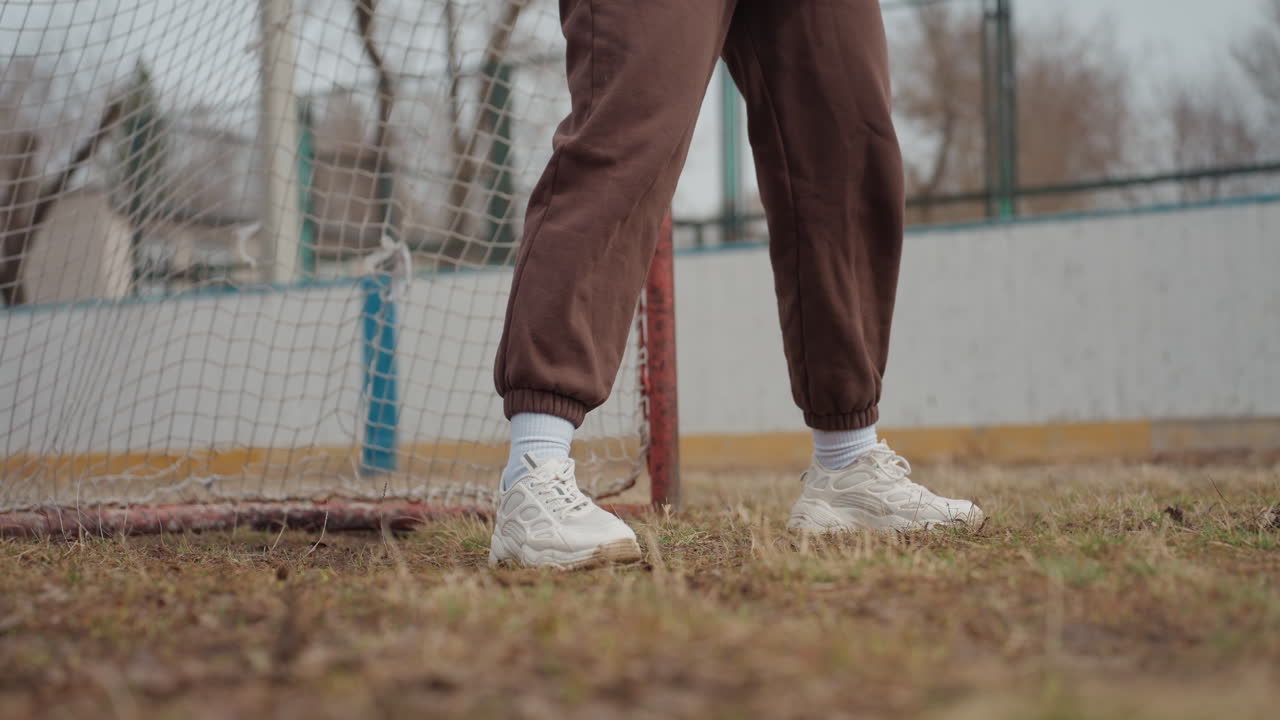 골 라인 근처에서 민첩하게 발놀림을 하는 백인 발과 운동화의 클로즈업, 발밑의 풀과 거친 흙, 갈색 캐주얼 스웨트팬츠와 흰색 양말이 보이고, 움직임에는 리듬감과 정확성이 느껴져요.