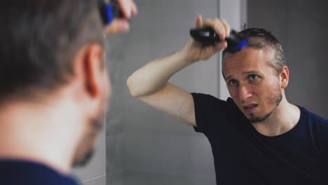 confused man shaves head using trimmer near large mirror