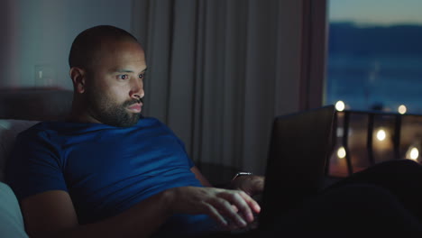 joven usando una computadora portátil acostado en la cama navegando en línea trabajando hasta tarde en la noche escribiendo mensajes de correo electrónico