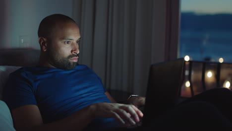 young man using laptop lying in bed browsing online working late at night typing email messages