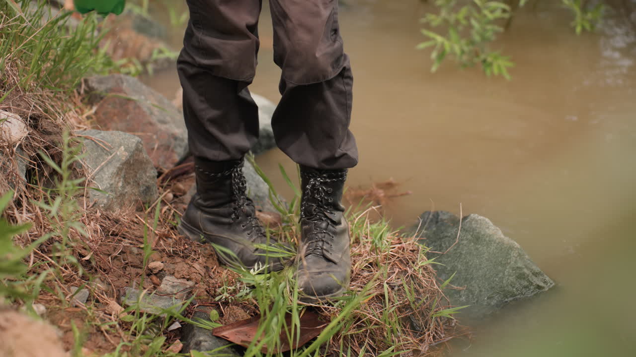 Back view of military man in tactical clothing squatting beside muddy riverbank while reaching into water, surrounded by overgrown vegetation, grass, stones, and damp soil during cloudy daylight