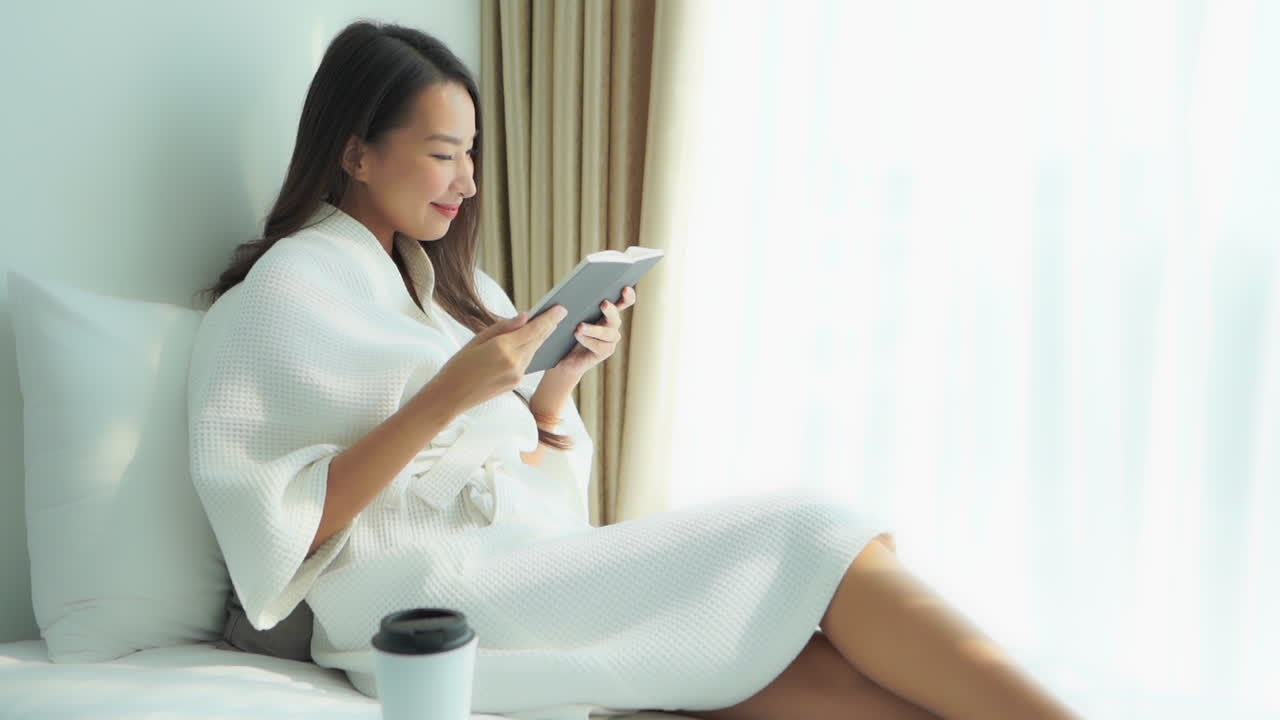 Attractive Chinese woman wearing white robe relaxing in bed with a book and cup of coffee. Bright sunlight through window on Asian lady smiling and laughing as she reads novel on vacation