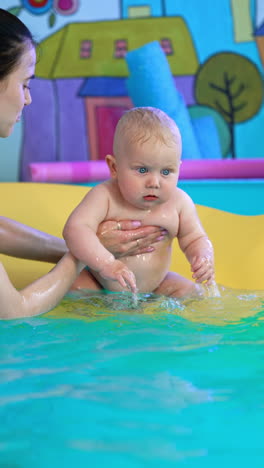adorable plump caucasian baby sits on the mat in the swimming pool. woman takes the child to her hands. vertical video.