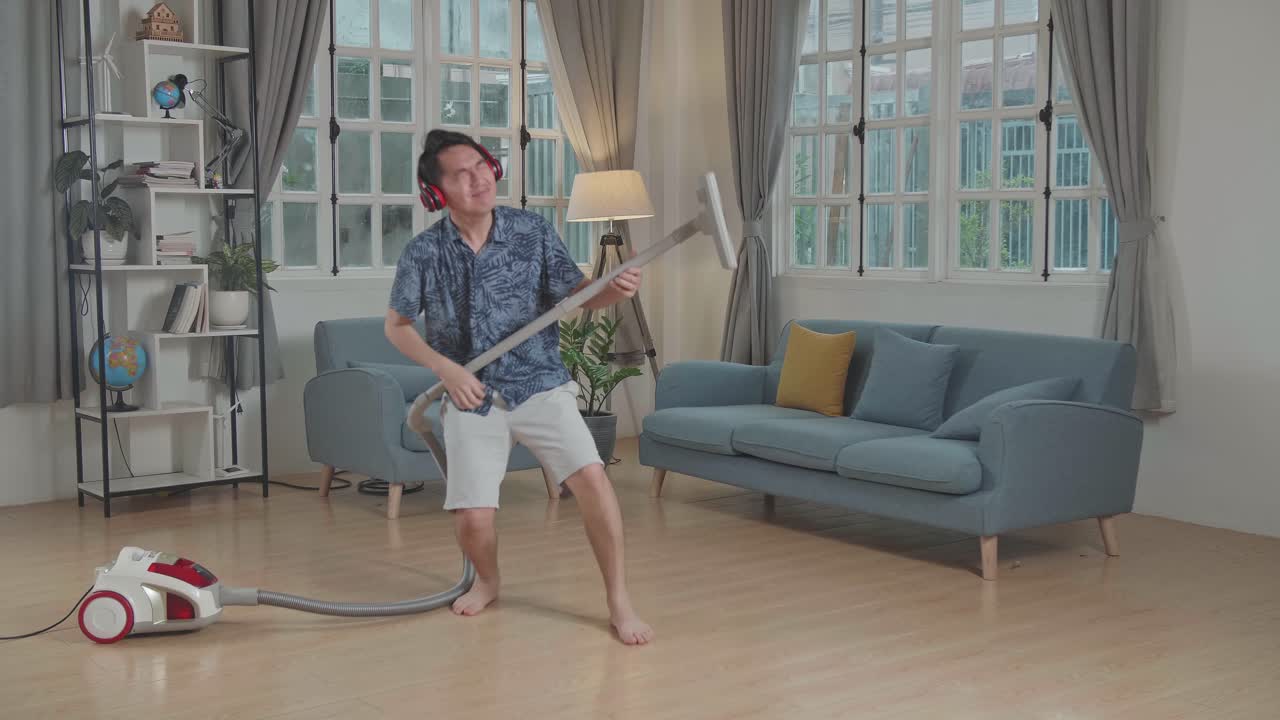 Cheerful Asian Man Cleaning Up His Home And Singing, He Is Using The Vacuum Cleaner As A Guitar And Microphone