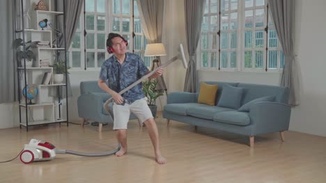 cheerful asian man cleaning up his home and singing, he is using the vacuum cleaner as a guitar and microphone