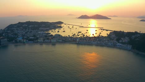 drone shot traveling backward from a city build on an isthmus with a long beach all along and an harbor during a beautiful sunset