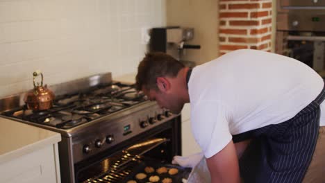 caucasian man wearing apron taking out tray of baked cookies from the oven in the kitchen at home