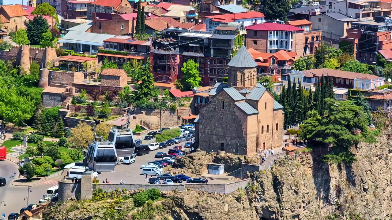 Funivie si muovono vicino a una chiesa storica e a edifici cittadini in una giornata di sole a Tbilisi, Georgia.