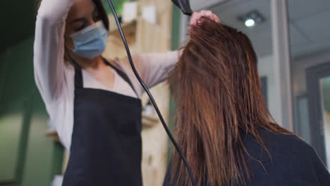 female hairdresser wearing face mask blow drying hair of female customer at hair salon