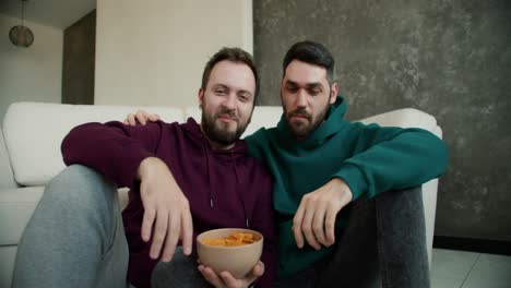 Gay-couple-enjoy-watch-TV-together-in-house,-discuss-and-eat-chips