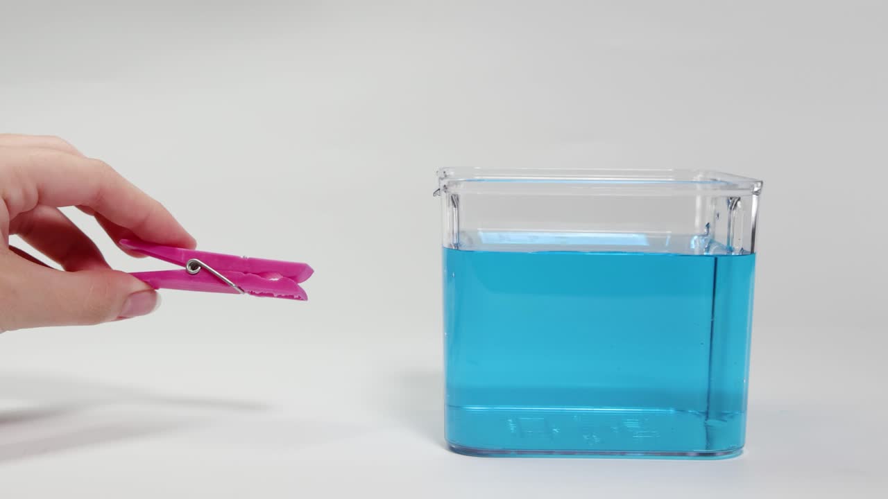 A hand drops a pink clothes peg into blue water, observing its buoyancy. Bright lighting, clear focus, scientific demonstration